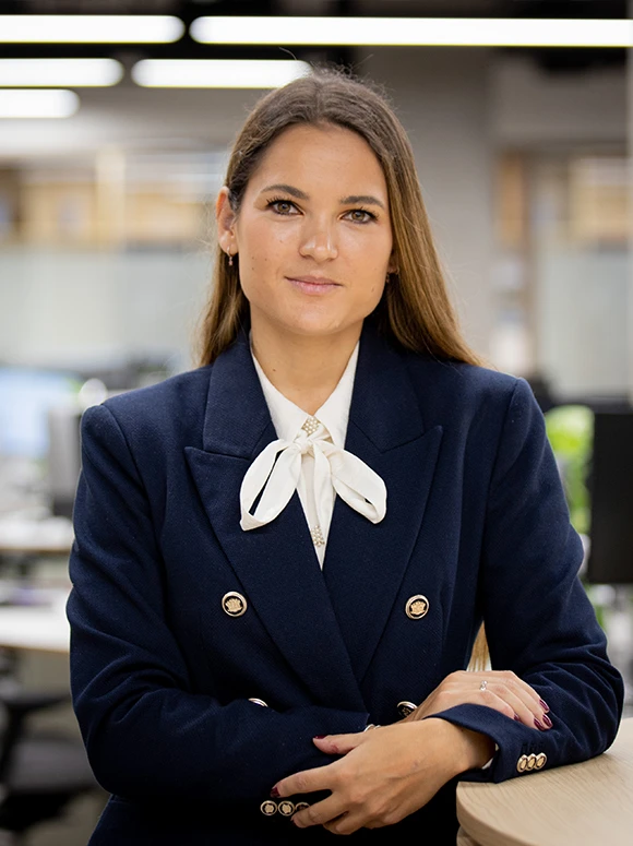 The image shows Sonia Gutierrez, a woman in a navy blue business suit with a white bow tie, posing for a professional headshot against a blurred background