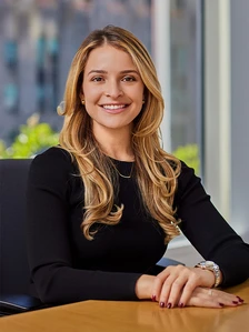 The image shows a smiling woman with long, wavy blonde hair wearing a black top, seated at a desk in an office environment