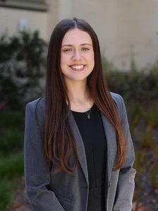 Malia Marshall, a young woman with long brown hair, smiles warmly while wearing a gray blazer and standing in front of some greenery