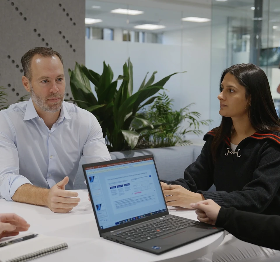 Two individuals, a man with a beard and a woman, discussing a laptop screen in an office setting with plants in the background