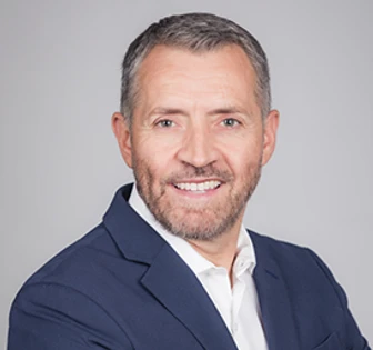 The image shows a headshot of Sy Pretorius, a smiling middle-aged man with gray hair wearing a navy blue suit and white collared shirt against a gray background
