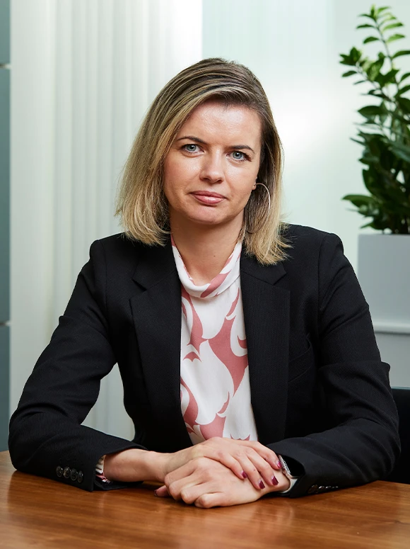 Michaela Rajnohova, a professional woman with blonde hair and blue eyes, sitting at a desk and wearing a black suit