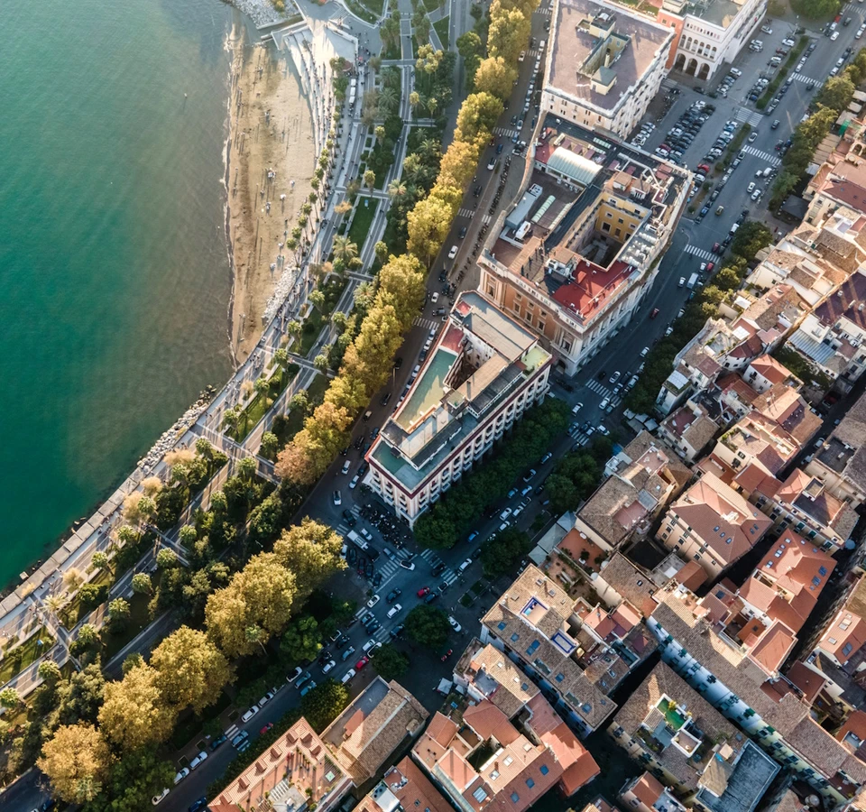 Aerial view of a vibrant city skyline with colorful buildings, a large body of water, and lush green trees lining the street