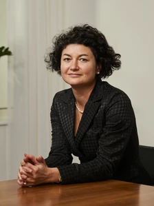 The image shows Nenka Berberiva, a middle-aged woman with curly dark hair, wearing a black patterned jacket, seated at a table with a serious expression