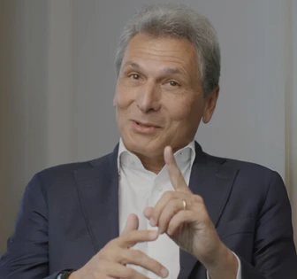 A mature, well-dressed man with short gray hair looks thoughtfully at the camera, his finger raised as if making a point