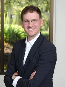 The image shows a smiling middle-aged man with dark hair and glasses, wearing a black suit and standing in front of a blurred nature background