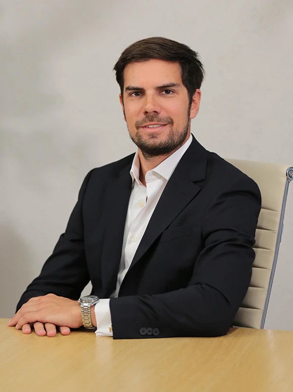 The image shows a well-dressed man with dark hair and a beard, sitting at a desk in what appears to be a professional setting. The filename suggests his name is Oscar Gomez Roldan