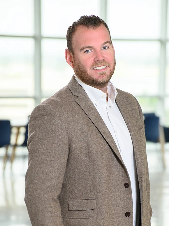 The image shows a smiling middle-aged man with a beard, wearing a gray tweed jacket, posing in front of a bright window