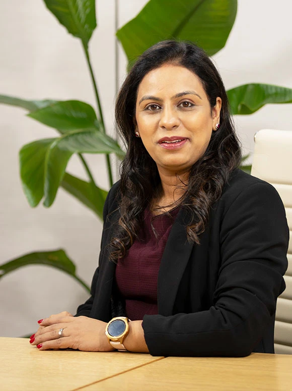 The image shows a seated woman with dark curly hair, wearing a black blazer and red top, sitting at a desk with green plants in the background. The filename indicates her name is Veenu Ahuja