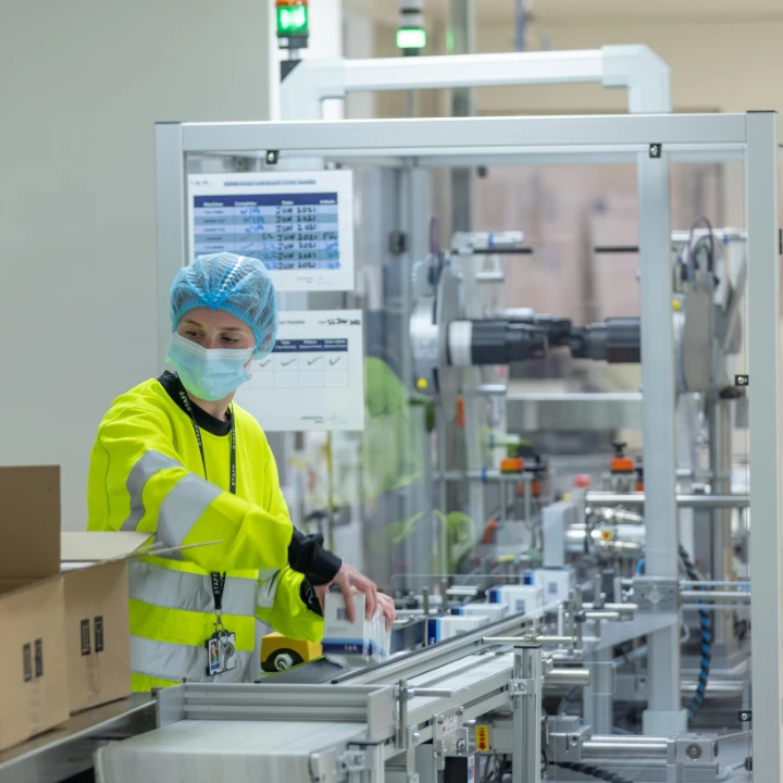 The image shows a person in a yellow safety vest and blue hairnet working at a pharmaceutical production facility, with various medical equipment and machinery visible in the background