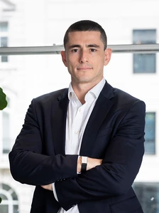 A professional-looking man in a suit stands with his arms crossed, appearing confident and focused in a modern office setting