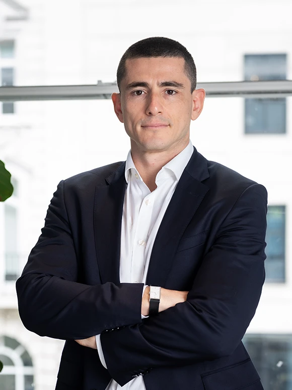 A professional-looking man in a suit stands with his arms crossed, appearing confident and focused in a modern office setting