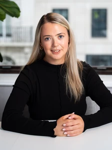 The image shows Judith Keane, a woman with long blond hair, wearing a black top, sitting at a desk and smiling at the camera