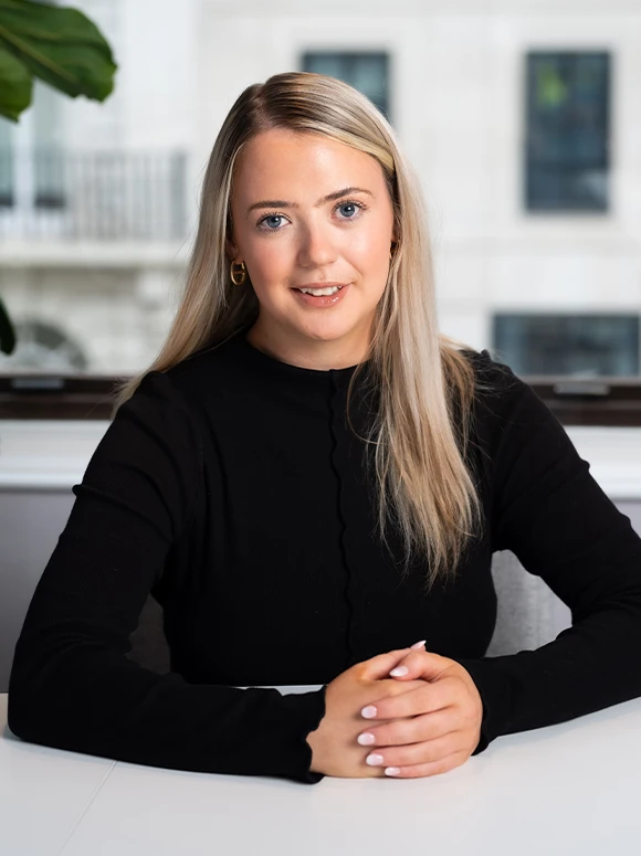 The image shows Judith Keane, a woman with long blond hair, wearing a black top, sitting at a desk and smiling at the camera