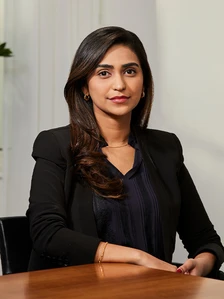 A professional-looking woman with long dark hair, wearing a black suit, sitting at a desk and looking directly at the camera