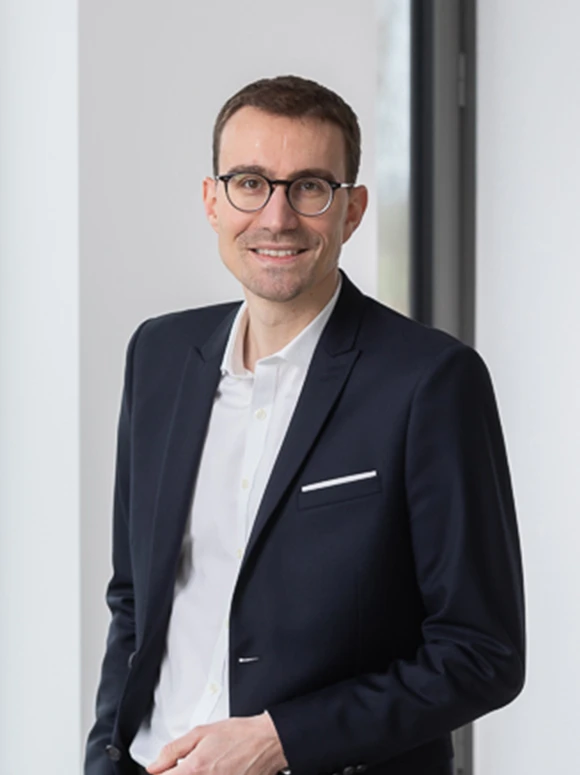 Headshot of a smiling, middle-aged man wearing a navy blue suit and glasses, standing in a professional setting