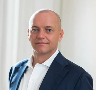 The image shows a close-up portrait of Francesco Pascalizi, a middle-aged man with blue eyes and a bald head, wearing a navy blue suit and white shirt against a neutral background