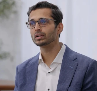 The image shows a man wearing glasses and a suit, with a thoughtful expression on his face, standing in an indoor setting