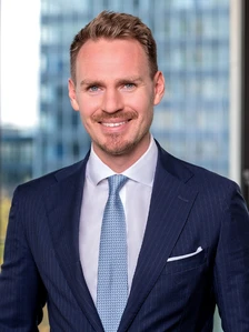 The image shows a smiling, well-dressed man in a navy blue suit and striped tie, with brown hair and a beard, standing in front of a blurred office building backdrop