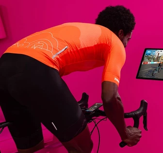A person in an orange jersey riding an exercise bike in front of a pink wall, with a tablet displaying a cycling activity on screen