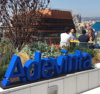 The image shows an outdoor seating area with large blue letters spelling "Adevinta" surrounded by lush greenery and people enjoying the space
